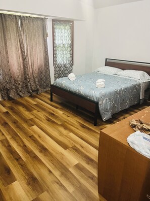 Ein Schlafzimmer mit Holzboden, einem Bett mit floralem Überwurf, einem Holz-Kopfteil und einem Vorhang mit geometrischem Muster.