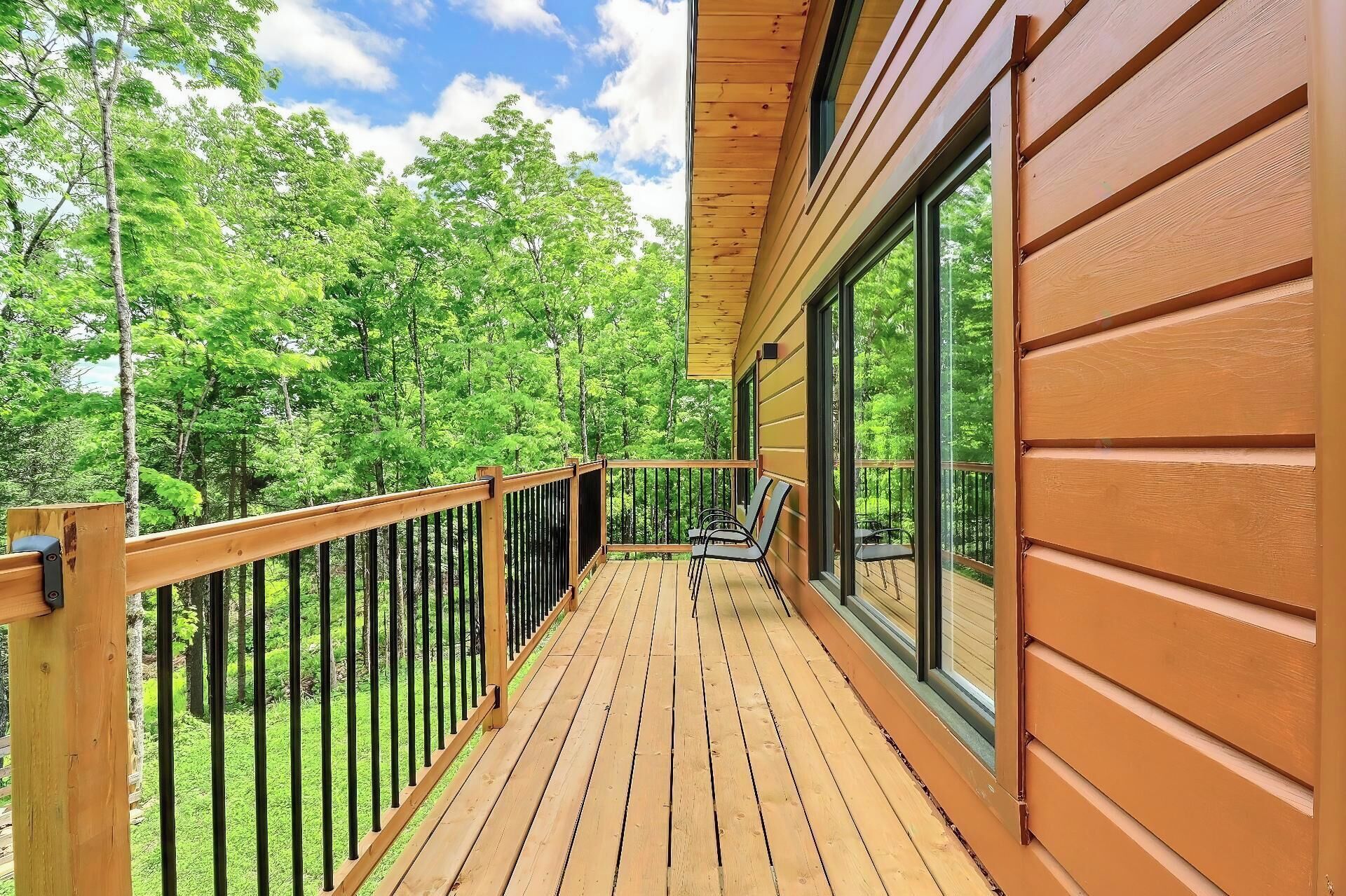 Une terrasse en bois avec une rambarde, de grandes fenêtres et une vue sur une forêt.