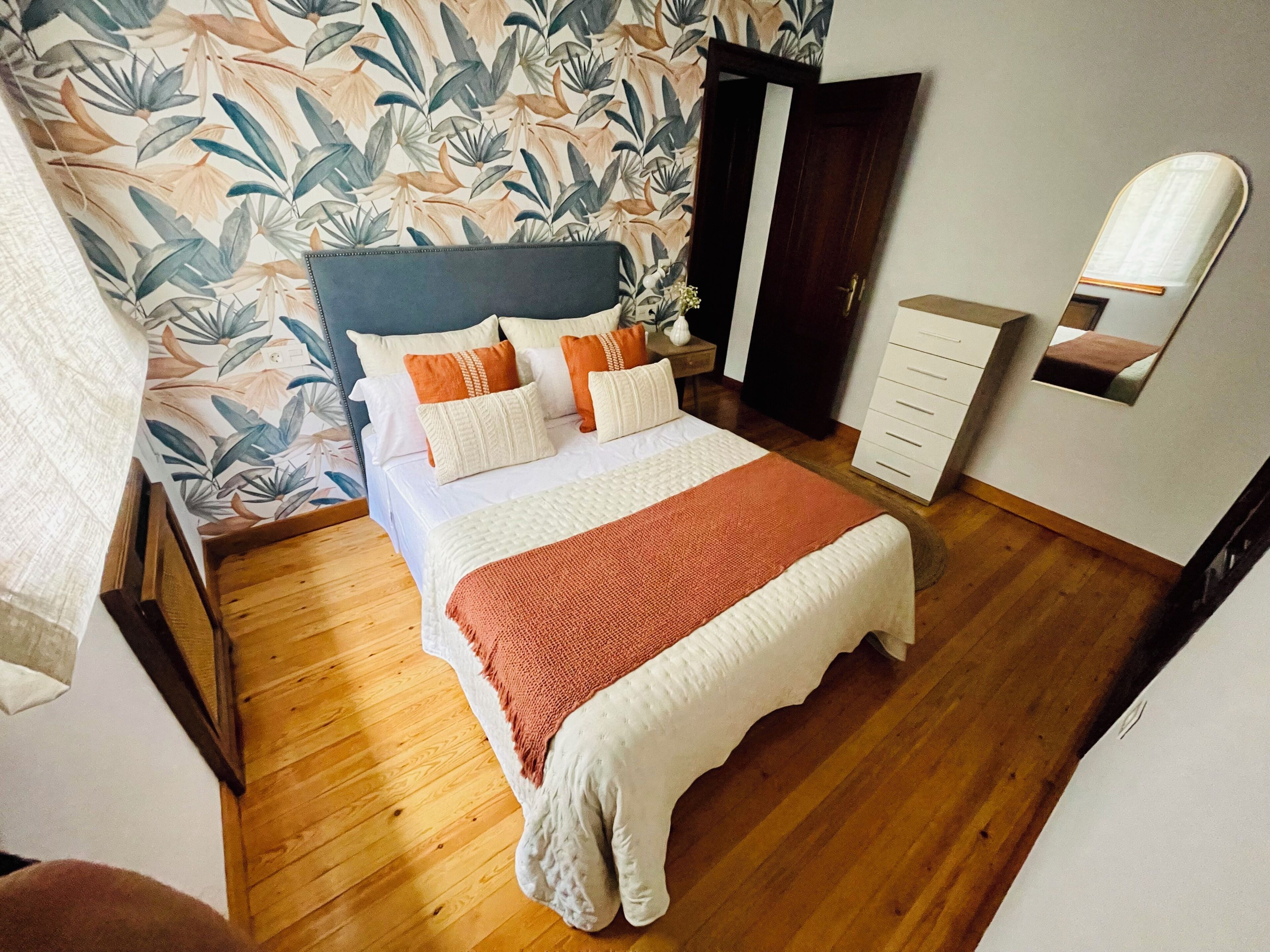 Une chambre à coucher comprenant un lit, une table de chevet, une commode, un miroir et un mur orné d’un motif de feuilles.