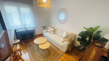 Una sala de estar con un sofá, dos mesas redondas, una mesa de comedor con sillas, una lámpara colgante, un espejo y una planta grande.
