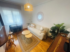 Ein Wohnzimmer mit einer Couch, zwei runden Tischen, einem Esstisch mit Stühlen, einer Pendelleuchte, einem Spiegel und einer großen Pflanze.