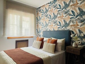 A bedroom with a bed, pillows, a bedside table with a lamp, a window with a sheer curtain, and a wall with a tropical leaf pattern.