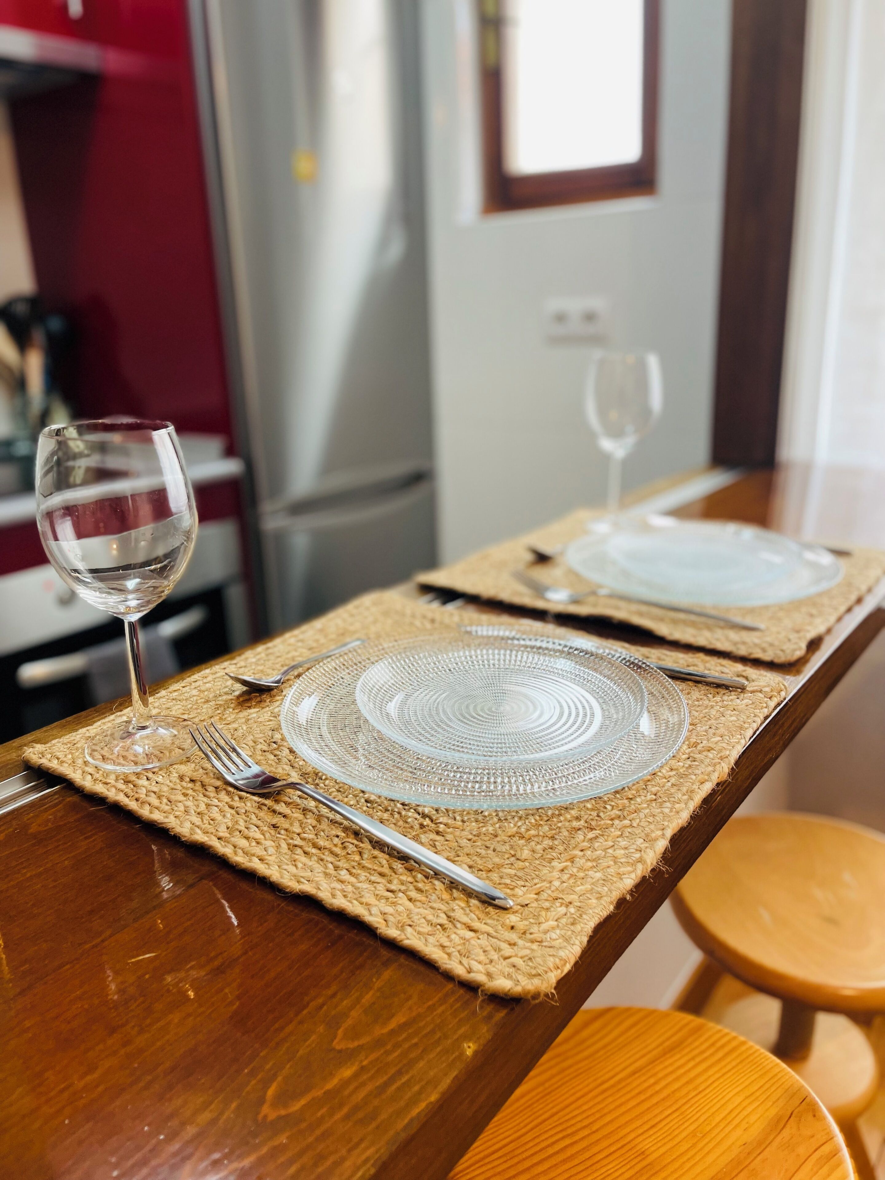 Una mesa con dos platos de vidrio transparente, un tenedor y una copa de vino sobre un mantel tejido.