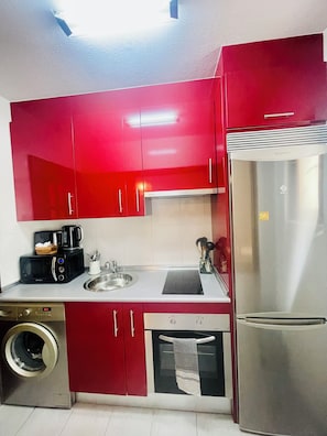 Eine kleine Küche mit roten Schränken, einem Edelstahl-Kühlschrank, einer Mikrowelle, einer Kaffeemaschine und einer Waschmaschine.