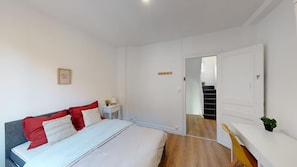 Een slaapkamer met een bed, een nachtkastje, een stoel, een kleine tafel, een potplant en een trap naar een andere kamer.