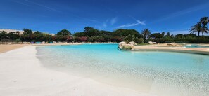 En klarblå pool med en sandstrand, omgiven av frodig grönska och palmer.
