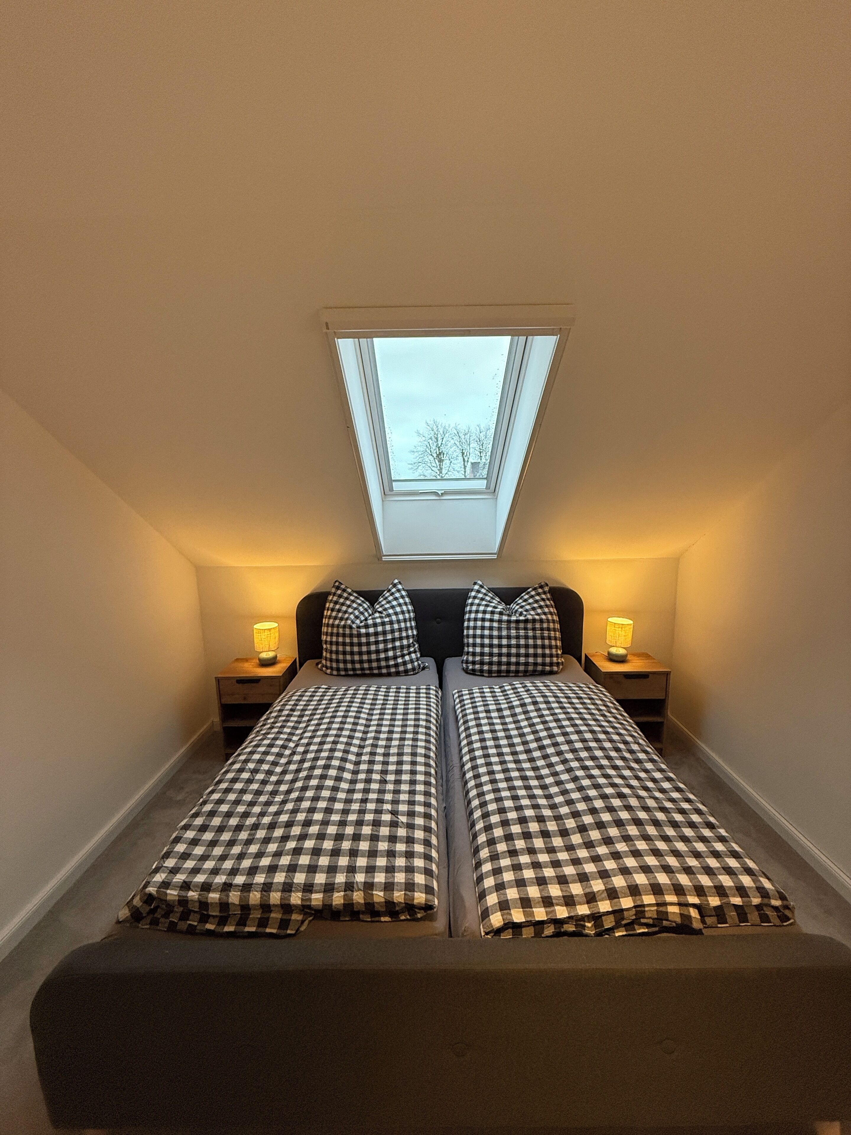 Ein Zimmer mit Dachfenster, zwei Betten mit karierten Bettbezügen, Nachttischlampen und einem dunklen Kopfteil.