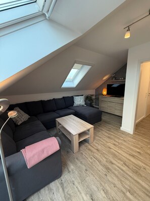 A modern loft apartment with a skylight, a sectional sofa, a wooden coffee table, and a TV in the background. - Vrbo Property (Wittmund)
