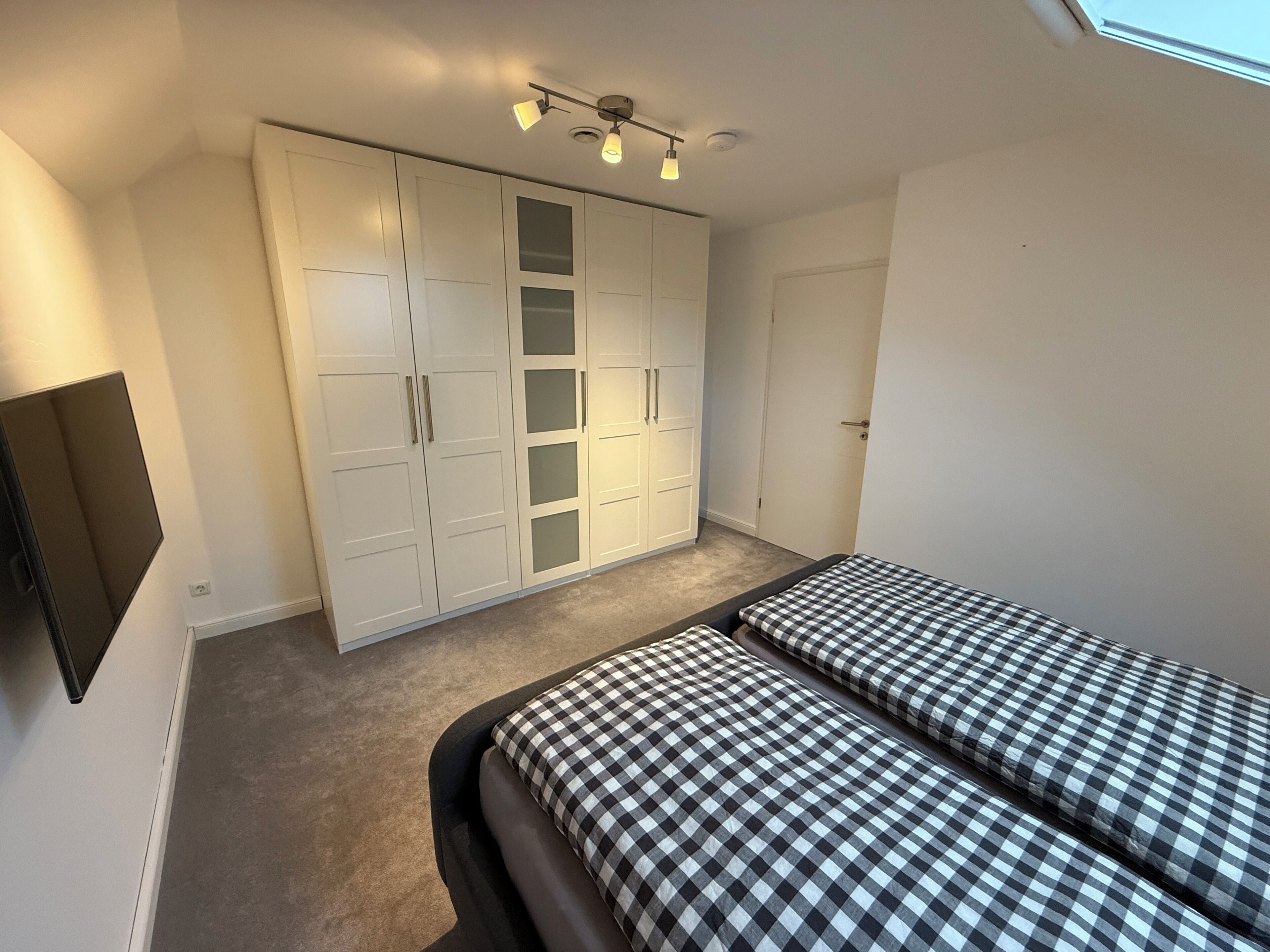 A bedroom with a large wardrobe, two beds with checkered bedding, a ceiling light fixture, and a wall-mounted TV.
