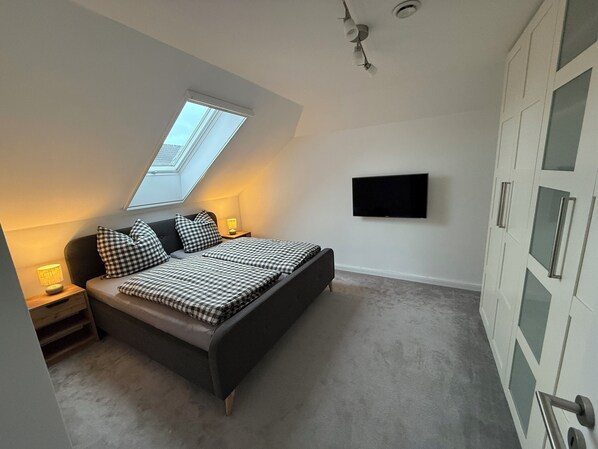 A bedroom with a bed, a skylight, a TV, a wardrobe, and a nightstand with a lamp. - Vrbo Property (Wittmund)