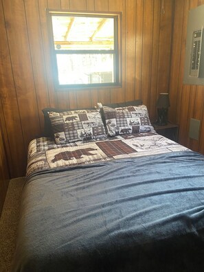 Ein Zimmer mit Holzwänden, einem Bett, einem Fenster und einem Nachttisch mit Lampe.