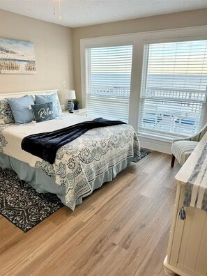 A bedroom with a large bed, two windows with blinds, a nightstand, a lamp, and a painting on the wall. - Beach Front - The Hamptons at Spanish Grant (Galveston)