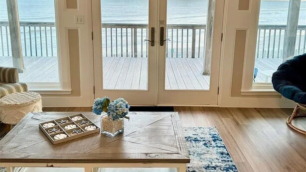 Ein Zimmer mit einer großen Glasdoor, die auf eine Holzterrasse mit Blick aufs Meer führt, einem kleinen Tisch mit Dekoration und einem blau-weißen Teppich.