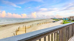 Blick auf einen Strand mit einer Holzlattenveranda, Liegestühlen und einer Reihe von Häusern.