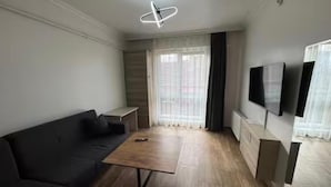 Una sala de estar moderna con un sofá oscuro, una mesa de centro de madera, un televisor de pantalla plana montado en la pared y un ventanal con cortinas de encaje.