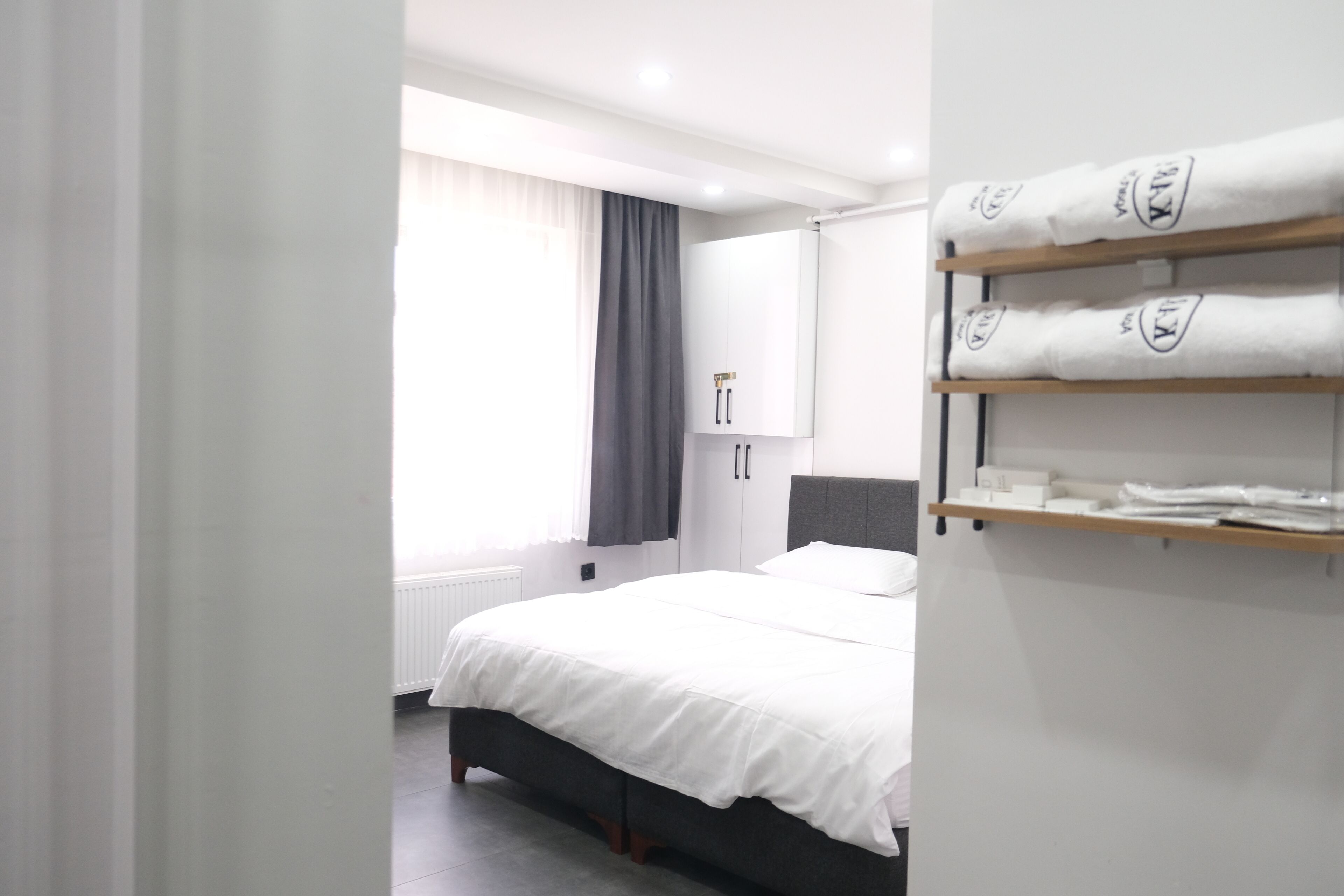 Una habitación de hotel con una cama, un armario, un estante con toallas y una ventana con cortinas.
