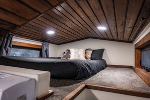 Un dormitorio con techo de madera, una cama con ropa de cama oscura, un almohadón con el texto 'HOME SWEET HOME' y una ventana con cortinas.