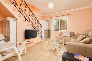 Una sala de estar con un sofá, una mesa de centro, un televisor, una silla y una escalera con barandilla decorativa.