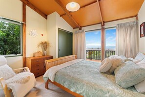 Quarto com cama, cadeira, cômoda, janela com cortinas e vista para o exterior.