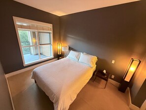 A bedroom with a large bed, a window with blinds, a bedside table with a lamp, and a nightstand with a lamp. - Resort-Style Condo In a Prestigious Golf Community (Victoria)