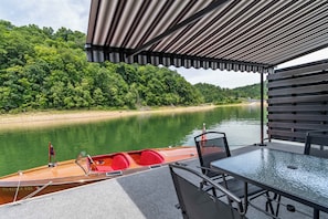 Un espace extérieur abrité, équipé d’une table et de chaises, avec un bateau amarré à proximité, et une rivière dont les berges sont bordées d’une colline boisée en arrière-plan.