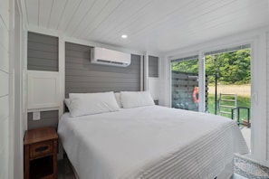 Un dormitorio con cama, ropa de cama blanca, una mesita de noche, un equipo de aire acondicionado y vistas al exterior.