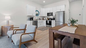 Une cuisine moderne avec des meubles blancs, un four à micro-ondes et un réfrigérateur. On y trouve deux chaises et une table en bois avec un set de table.