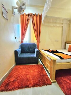 A bedroom with a bed, a chair, a fan, and a window with curtains. - angel motel ntinda (Kampala)