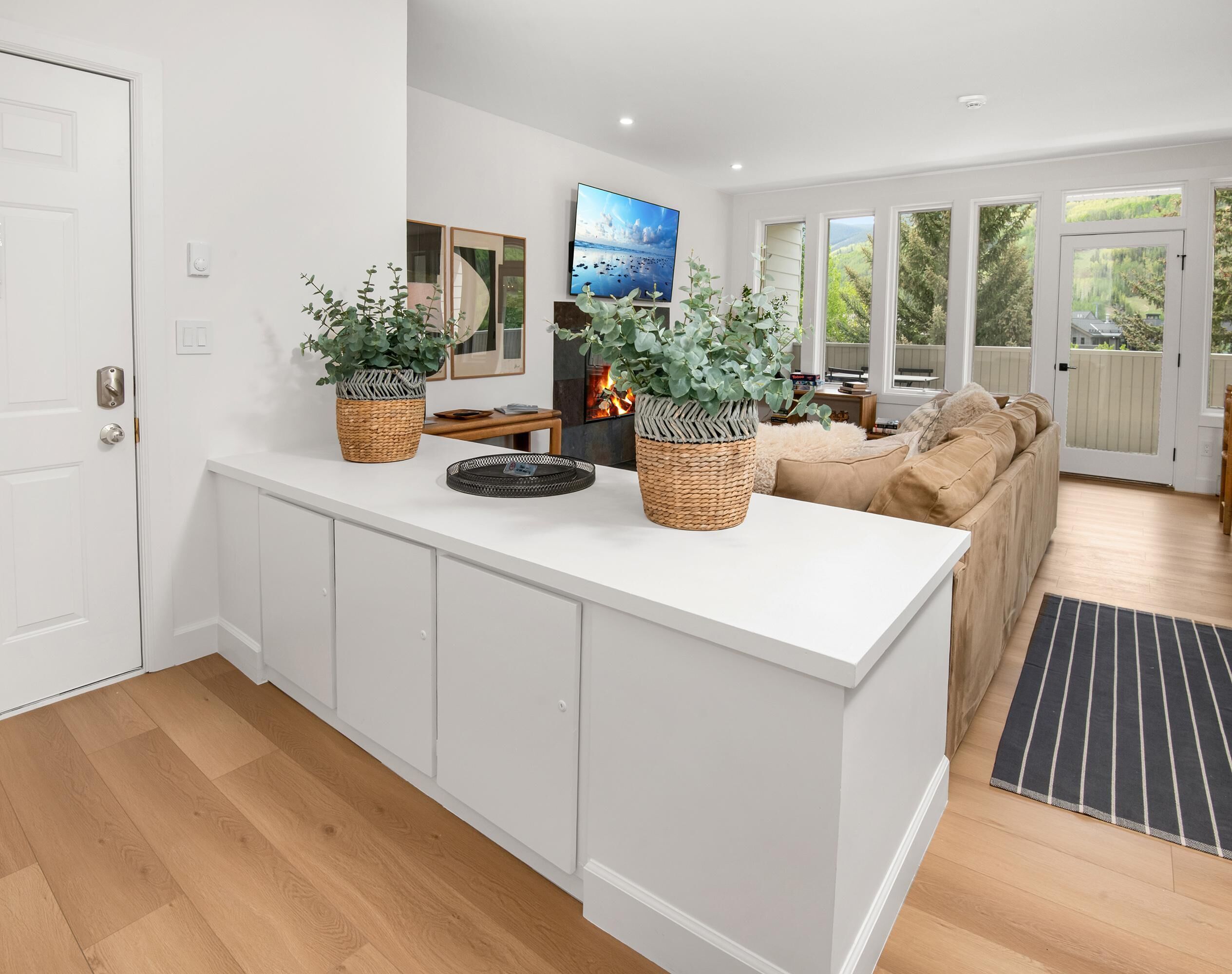 A modern kitchen with white cabinetry, a central island with wicker baskets, a flat-screen TV, and a cozy seating area with a sofa and a fireplace.