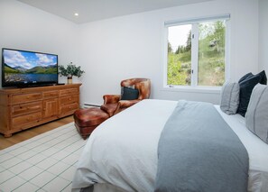Ein Schlafzimmer mit einem Bett, einem Ledersessel, einem Fernseher auf einer Holzkommode und einem Fenster mit Blick auf Grünflächen.