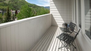 Un balcón con vistas a una montaña boscosa, barandilla blanca y una silla plegable de metal.