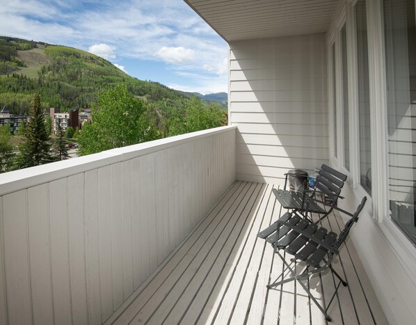 Ein Balkon mit Blick auf einen bewaldeten Berg, einem weißen Geländer und einem klappbaren Metallstuhl.