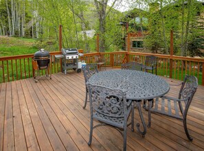 Eine Holzterrasse mit einem runden Metalltisch und Stühlen, einem Grill und einer Steinstruktur im Hintergrund.