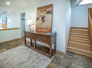 Une pièce avec un escalier en bois, une affiche représentant Ecker Hill, une console en bois avec une lampe et un tapis à motifs.