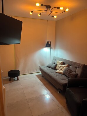 A living room with a sofa, a small ottoman, a floor lamp, and a flat-screen TV mounted on the wall. - Apartamento completo, próximo ao aeroporto de Guarulhos - Via alameda (Guarulhos)
