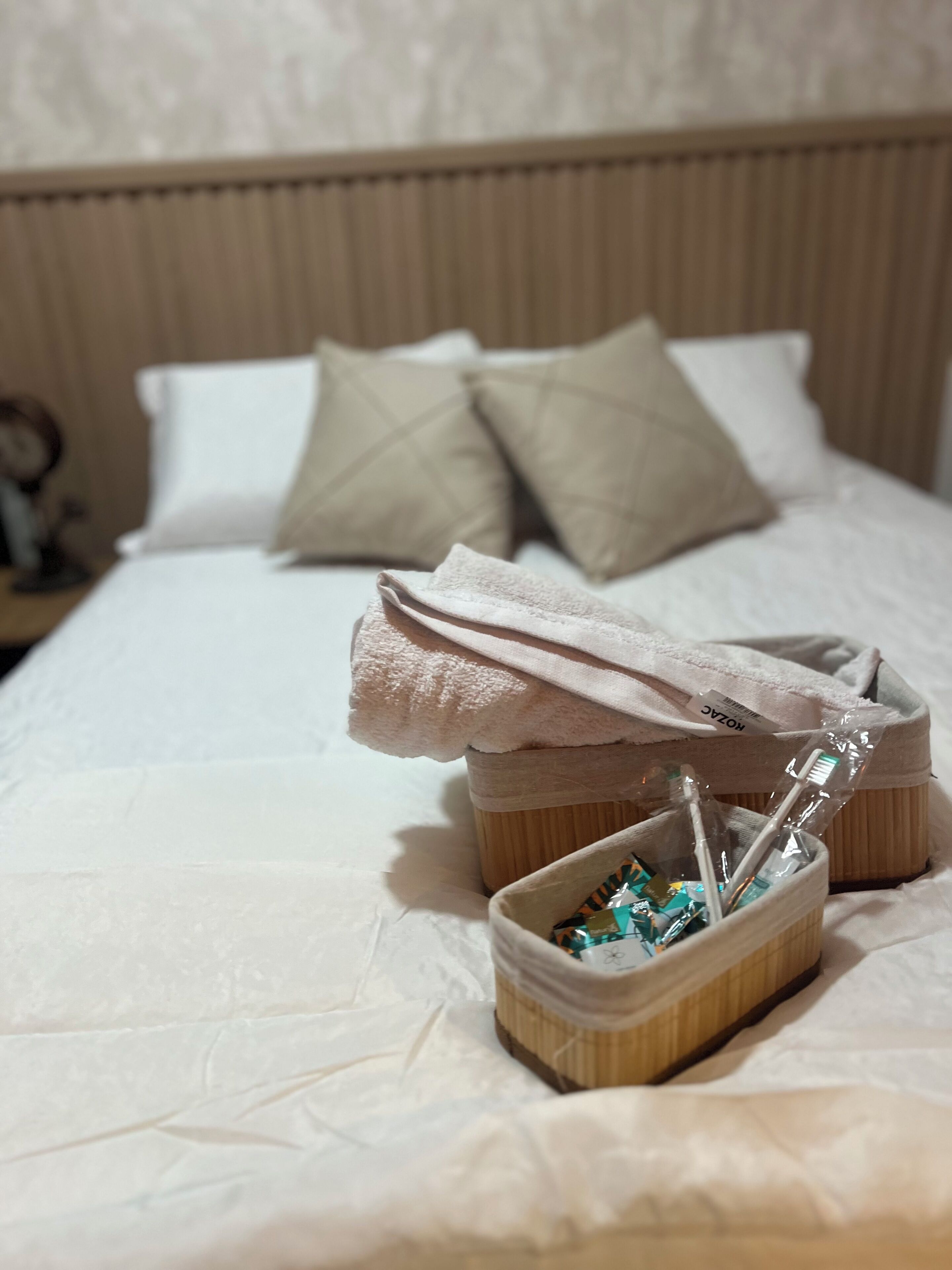 A neatly made bed with pillows, a small wooden basket with toiletries, and a folded towel on the bed.