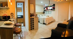 A modern living room with a sofa, a TV mounted on the wall, and a kitchen area with a dining table and chairs. - Apartamento completo, próximo ao aeroporto de Guarulhos - Via alameda (Guarulhos)