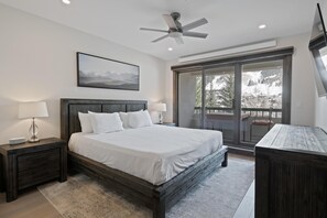 Ein Schlafzimmer mit einem großen Bett, einem Deckenventilator, einem Nachttisch mit Lampe, einem Schreibtisch und Blick auf eine verschneite Landschaft durch eine Schiebetür.