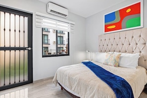 Une chambre à coucher avec un lit, une fenêtre avec des stores, un appareil de climatisation et un tableau abstrait aux couleurs vives accroché au mur.