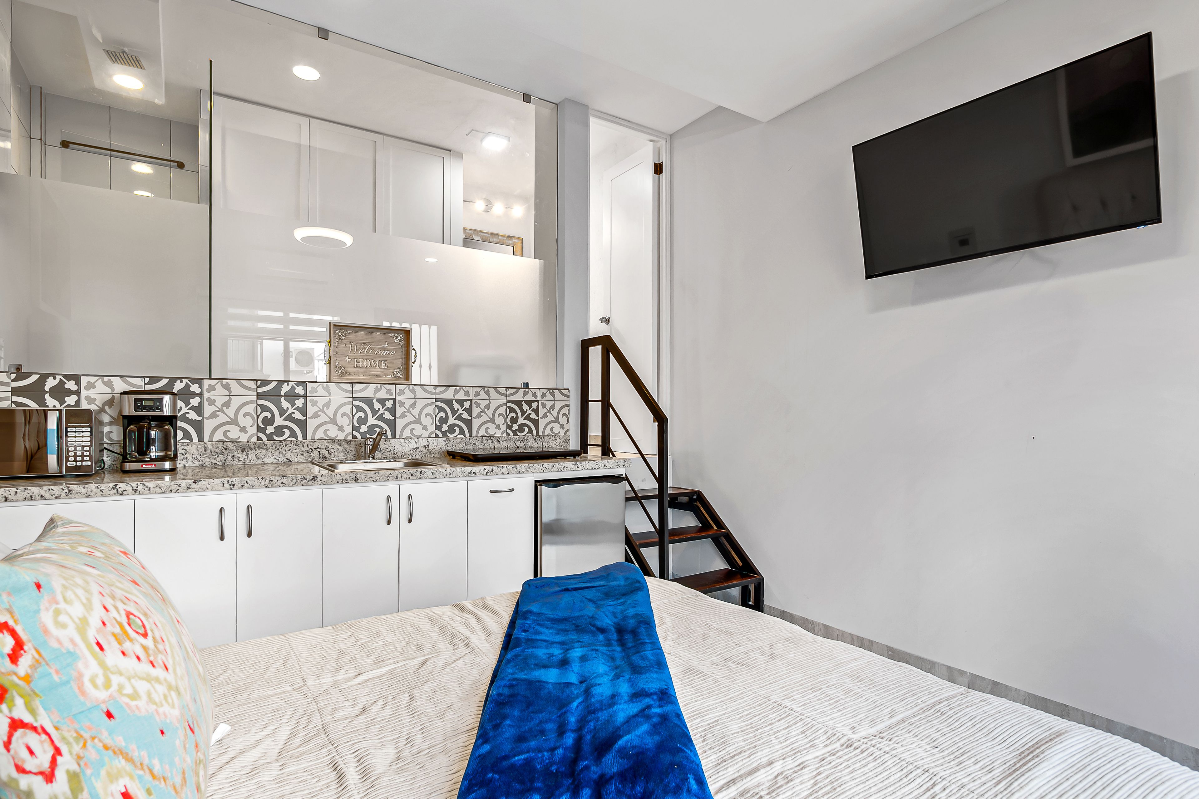 A modern hotel room with a bed, a blue blanket, a TV mounted on the wall, a kitchen area with white cabinets, and a tiled backsplash.