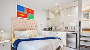A modern bedroom with a tufted headboard, a bed with a blue blanket, a nightstand, and a small bedside table. Adjacent to the bedroom is a kitchenette with a refrigerator, a microwave, and a coffee maker. - Morgan Residences 603 at Boutique Hotel With Pool, Gym & Spa (Cabo San Lucas)