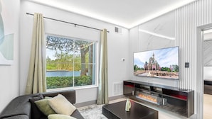 Ein modernes Wohnzimmer mit großem Fernseher, Kamin und Blick auf ein Schloss durch das Fenster.