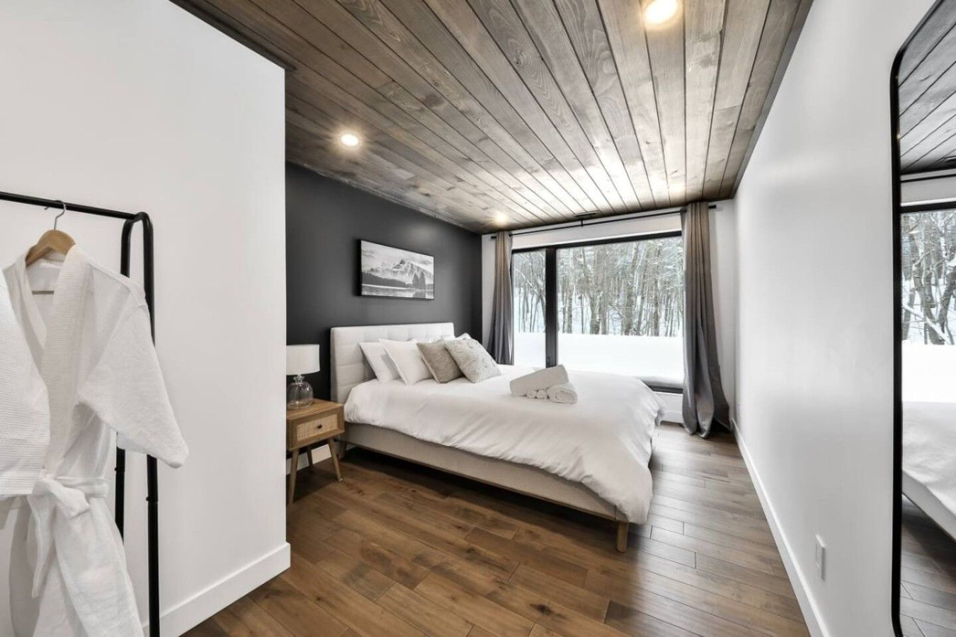 A modern bedroom with a wooden ceiling, a large bed, a nightstand, and a window with a view of trees.
