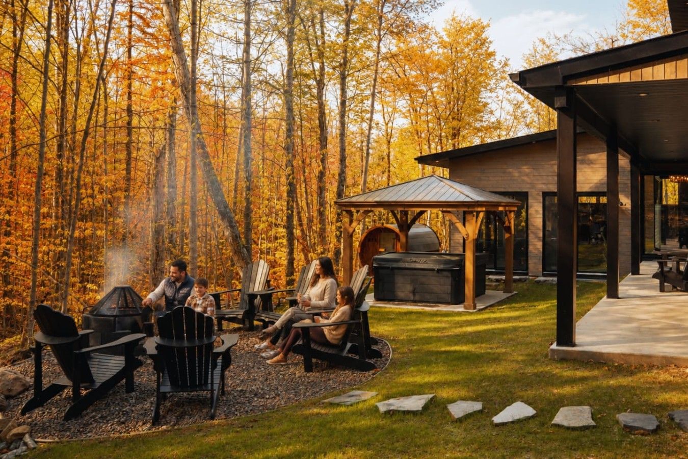 A family gathered around a wood-fired grill in a forested outdoor area with a covered seating area and a sauna.