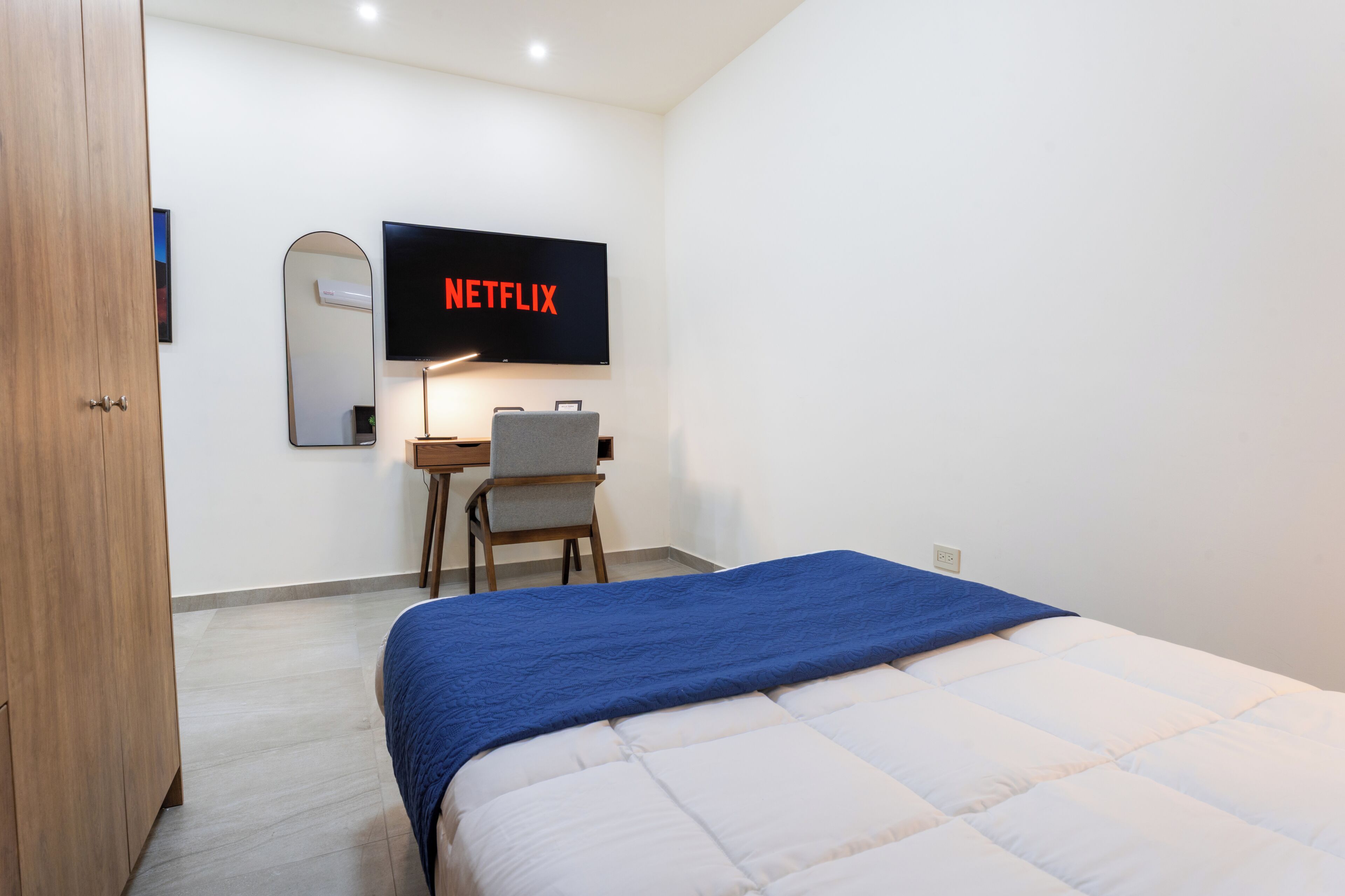 A bedroom with a bed, a desk, a chair, a TV mounted on the wall, and a wooden wardrobe.