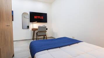 A bedroom with a bed, a desk, a chair, a TV mounted on the wall, and a wooden wardrobe.