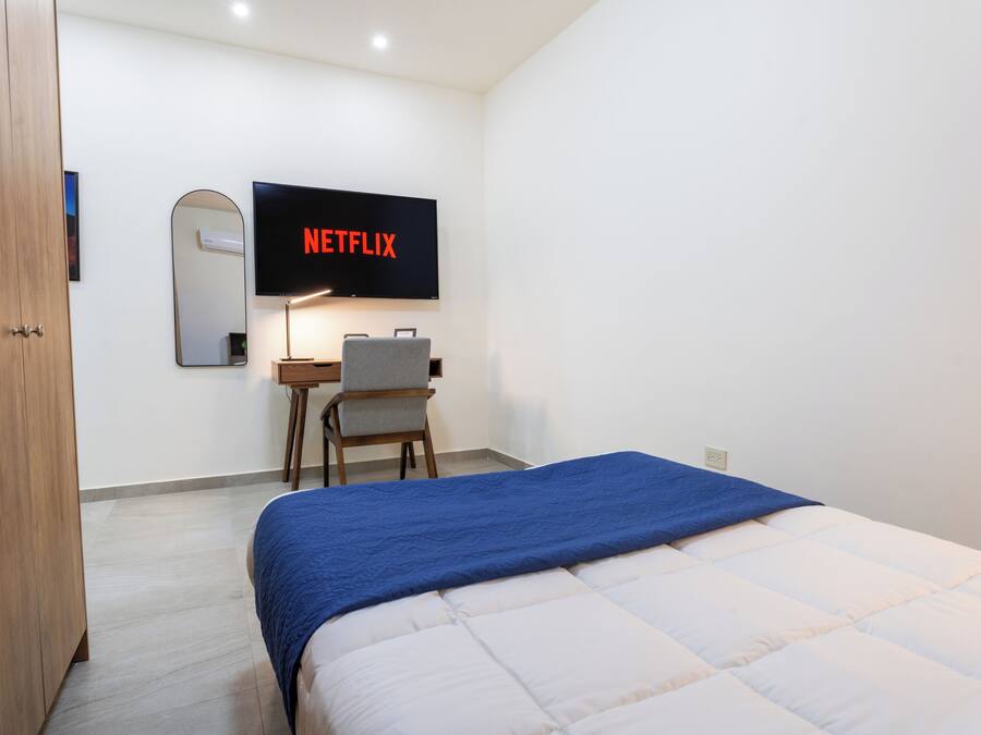 A bedroom with a bed, a desk, a chair, a TV mounted on the wall, and a wooden wardrobe.