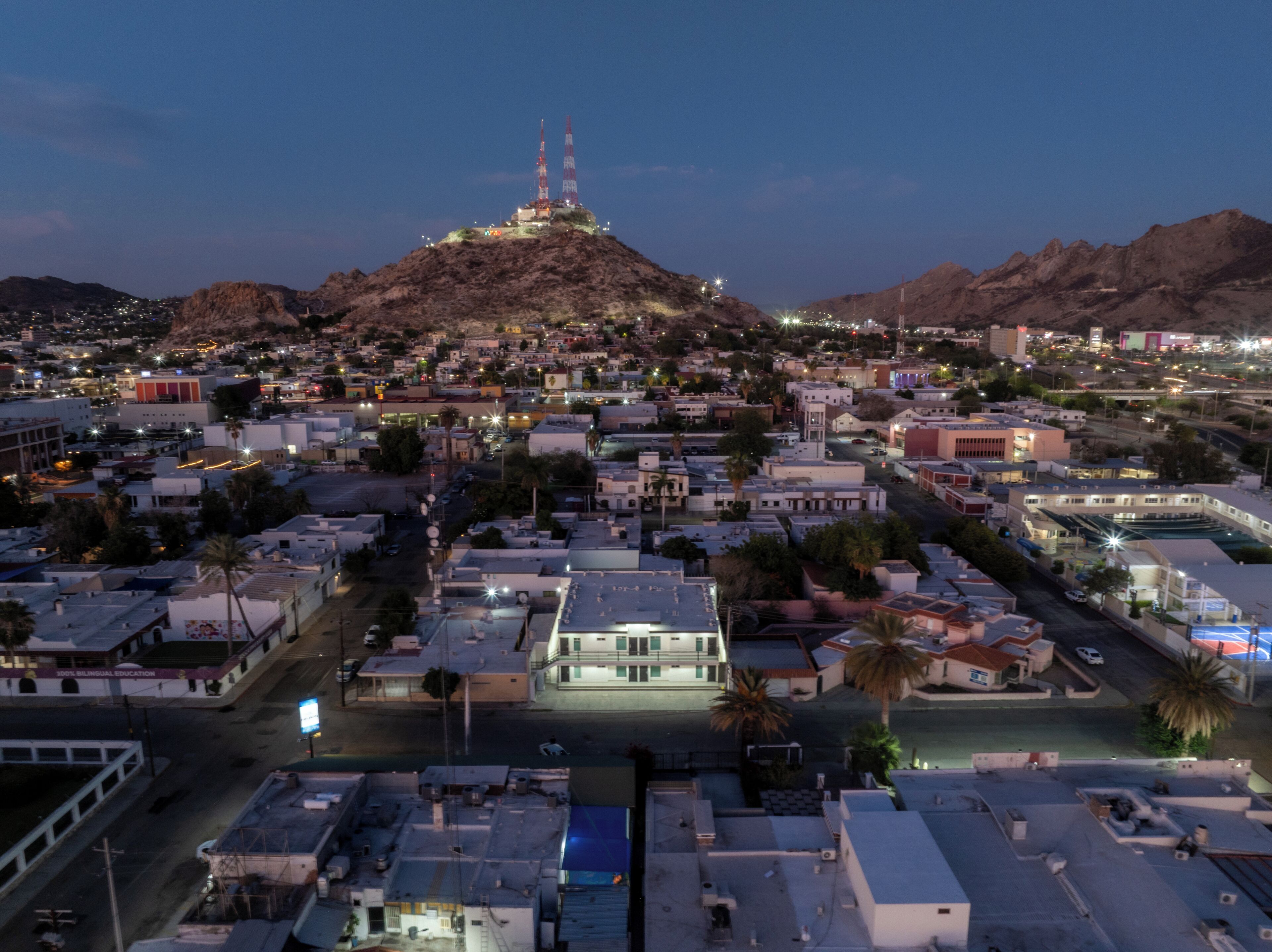 Un paisaje urbano con una estructura destacada en la cima de una colina, posiblemente una torre de comunicaciones, rodeada de edificios residenciales y calles.