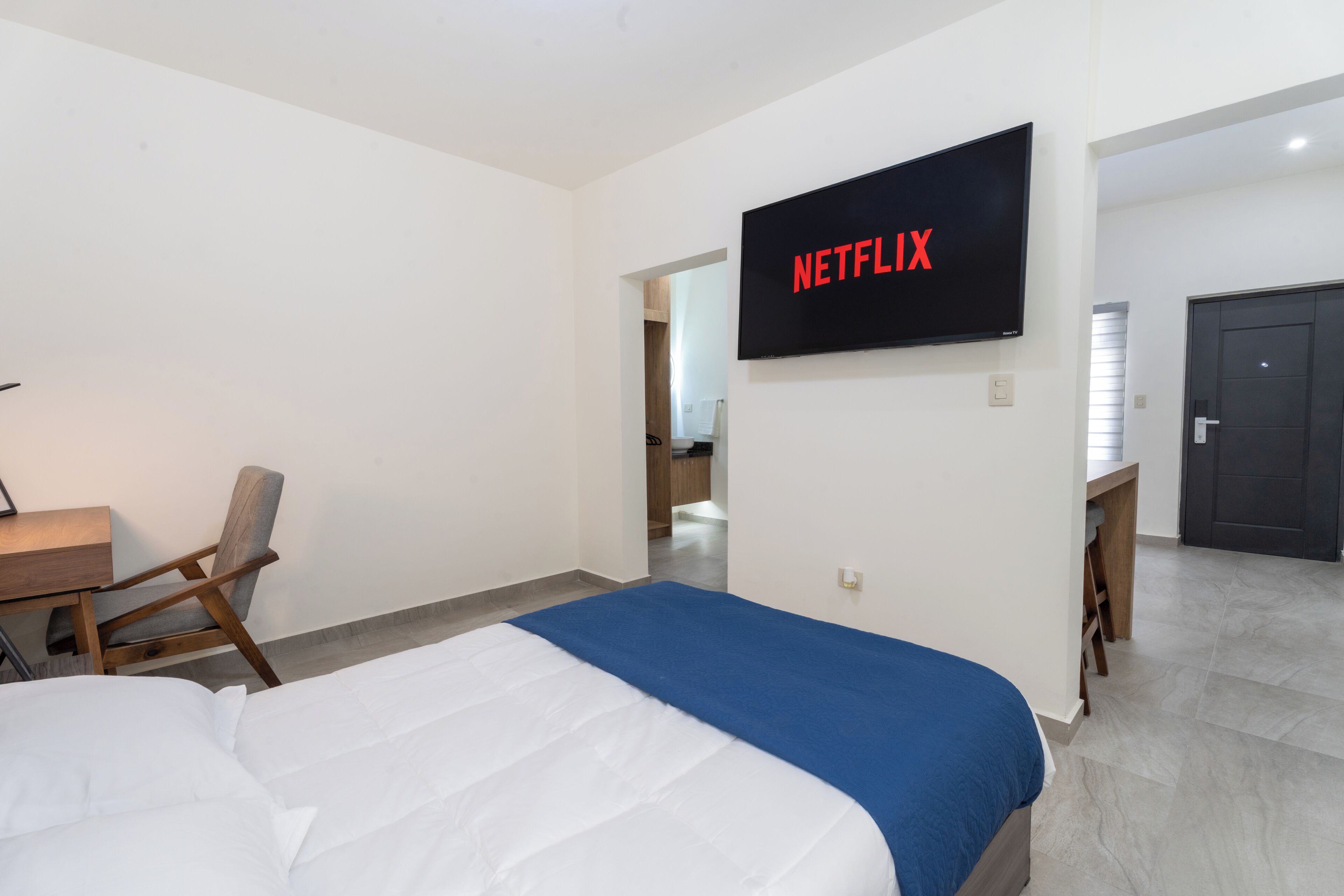 A hotel room with a bed, a desk, a chair, a TV mounted on the wall, and a door leading to another room.