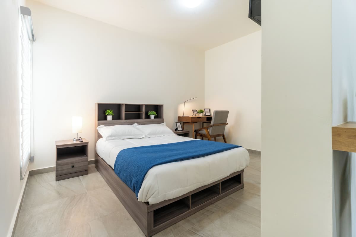 Une chambre à coucher comprenant un lit, une table de chevet, une lampe, un bureau, une chaise et une petite plante.